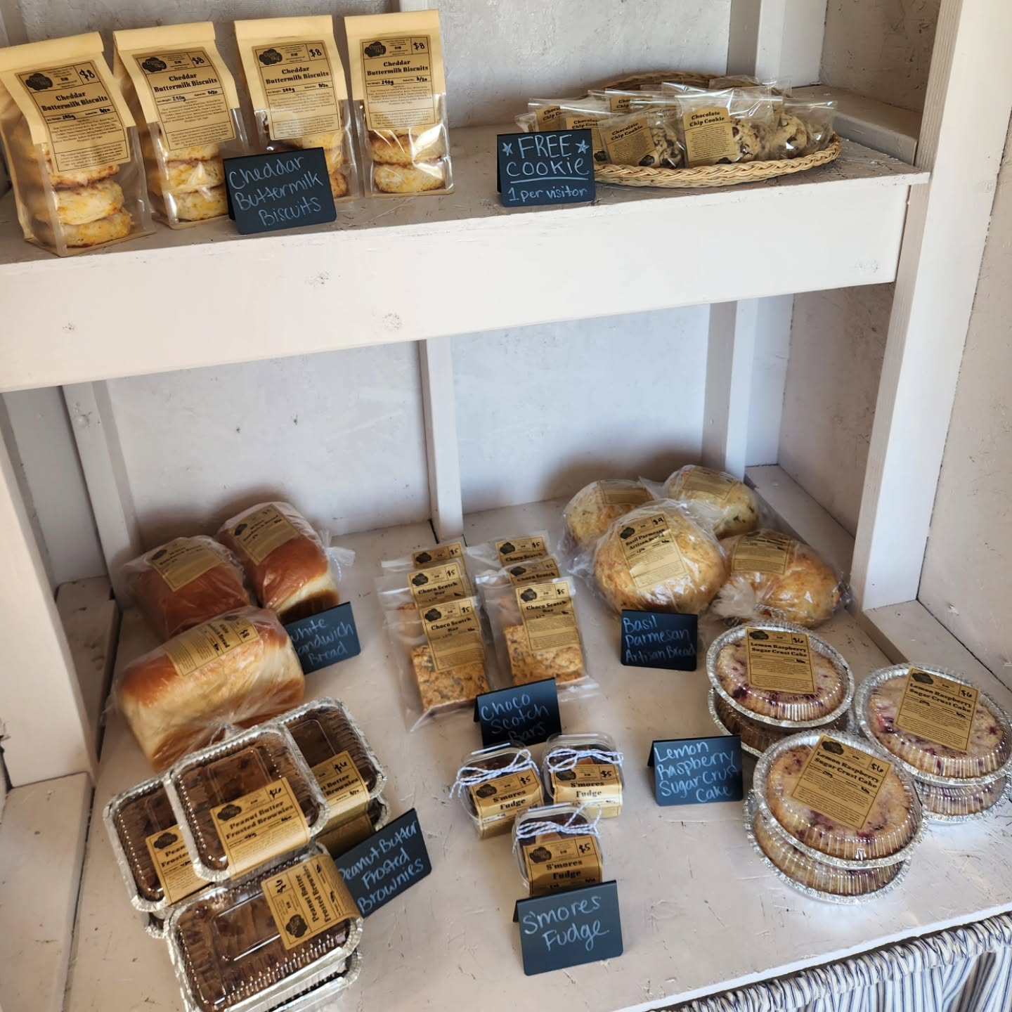 White self-serve shelves stocked with labeled breads, biscuits, brownies, fudge, and lemon raspberry cakes, plus a free cookie basket